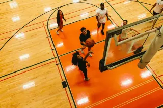 Birds-eye view of two students playing basketball on an indoor court with orange flooring. One student, a young woman, shoots the basketball while another student, a young man, runs beside her. Both are wearing black clothing and athletic shoes. Court markings and part of a basketball hoop are visible in the frame.
