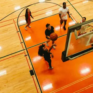 Birds-eye view of two students playing basketball on an indoor court with orange flooring. One student, a young woman, shoots the basketball while another student, a young man, runs beside her. Both are wearing black clothing and athletic shoes. Court markings and part of a basketball hoop are visible in the frame.