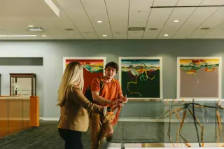 A Sam Houston student in an orange polo talks with a blonde professor in a blazer. They are standing in an art gallery with two large, framed pieces of art behind them.