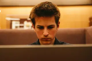 A young man with short brown hair is deeply focused on working on a laptop. He is seated in a quiet area with warm ambient lighting. The background features wooden paneling and blurred shelves, creating a studious atmosphere.