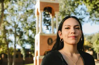 A young woman looks at the camera, smiling. Behind her is the Sam Houston State University clock tower and branches from several trees.