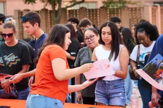 A Sam Houston student in an orange shirt stands behind an orange table outside on campus, showing a brochure to another student in a white tank top and shorts. There are many other students in the background looking for information from other tables.