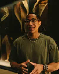 A young man wearing glasses, a green t-shirt, gold chain necklace, and a watch, smiling warmly and gesturing with his hands. The background is softly blurred with earthy tones.