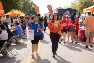 The camera focuses on two students, both young women, walking towards it amidst a busy outdoor event. One student wears a bright blue shirt and black shorts, while the other is in an orange shirt and black pants; they are holding various drink cans and food. In the background there are people walking around and sitting amongst pop-up tents and food trucks. Bright sunlight and greenery in the distance enhance the lively and festive ambiance.
